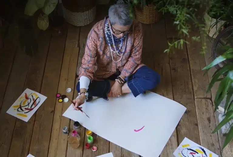 Mujer mayor sentada en el suelo, rodeada de materiales de pintura y papeles, creando una obra de arte. El espacio está decorado con plantas, transmitiendo una atmósfera relajada y acogedora, que refleja las oportunidades económicas del envejecimiento demográfico activo y productivo.