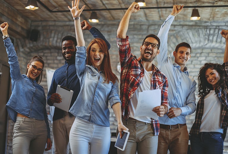Grupo diverso de jóvenes sonrientes celebra con los brazos en alto en un espacio de oficina moderno con paredes de ladrillo y tonos cálidos. La escena transmite entusiasmo y trabajo en equipo dentro de una acción social centrada en la inclusión laboral y juvenil.