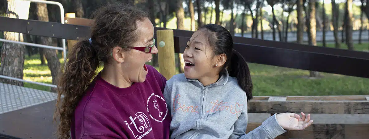 Una mujer con gafas negras, pelo recogido en una coleta y sudadera burdeos comparte un momento alegre con una niña sonriente en sudadera gris, en un parque rodeado de árboles. Tonos cálidos y naturales resaltan esta escena de acción social que transmite inclusión, empatía y conexión humana.