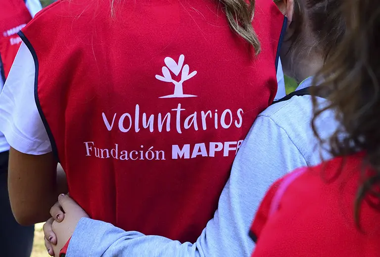 People embraced from behind wear red volunteer vests from Fundación MAPFRE, with a white logo on a vibrant background. The image conveys unity and commitment in a collaborative social action that promotes volunteering, solidarity, and community support in diverse contexts.