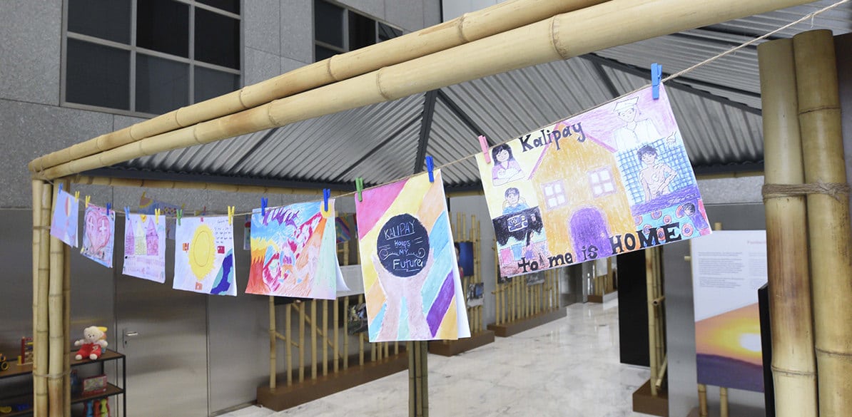 Exposiciones sociales Dibujos infantiles coloridos cuelgan con pinzas sobre cuerda entre estructuras de bambú en una exposición interior. Tonos vivos y mensajes positivos destacan una acción social centrada en la infancia, creatividad y expresión artística como herramienta de inclusión y desarrollo emocional.