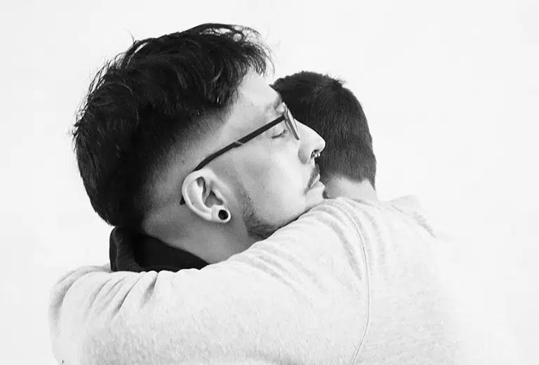 Salud mental Dos personas se abrazan con fuerza en una imagen en blanco y negro, transmitiendo empatía y apoyo emocional. El fondo neutro y los tonos grises refuerzan el mensaje de una acción social centrada en la salud mental, el acompañamiento y la reconciliación.