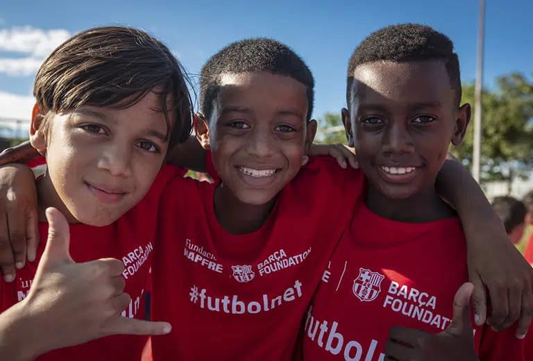 Futbolnet: cuando el deporte no es sólo un juego Tres niños sonrientes con camisetas rojas de Fundación Barça y Fundación MAPFRE se abrazan en una cancha deportiva al aire libre. El cielo azul y la luz natural enmarcan una acción social que fomenta la inclusión, el deporte y la amistad infantil.