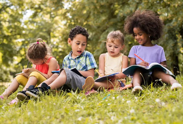 Cuatro niños pequeños sentados sobre el césped, dibujan y escriben al aire libre en un entorno natural y soleado. Con ropa colorida y expresiones concentradas, la escena refleja una acción social educativa que promueve la creatividad, inclusión y aprendizaje en la infancia.