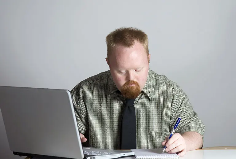 Hombre con síndrome de Down, camisa a cuadros y corbata negra, trabaja concentrado en su computadora portátil y libreta. Fondo claro y tonos neutros en una escena que refleja inclusión laboral dentro de una acción social orientada a la igualdad de oportunidades.