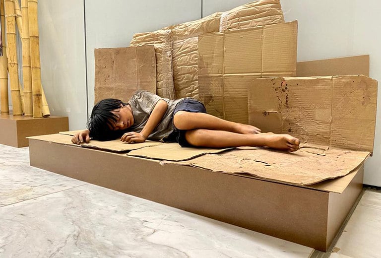 Kalipay: un viaje a la esperanza Niño descalzo y con ropa sucia yace dormido sobre una estructura de cartón que simula un banco, en una instalación artística. Tonos tierra refuerzan el mensaje de una acción social que denuncia la pobreza infantil y la exclusión habitacional.