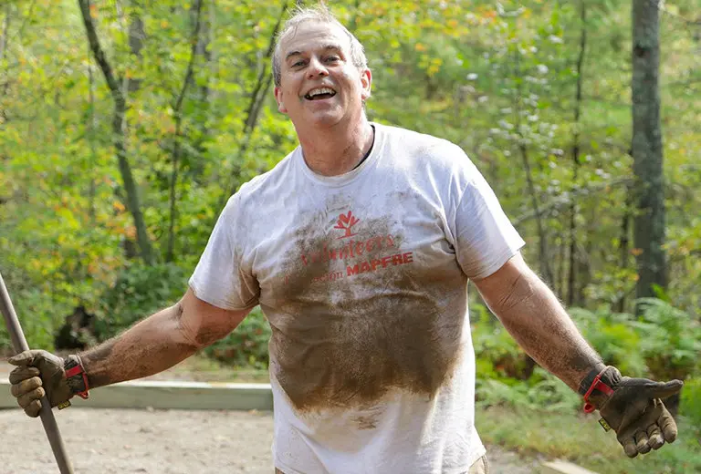 Voluntario sonriente con camiseta blanca manchada de tierra trabaja al aire libre en un entorno boscoso. Con guantes y herramientas, la escena refleja una acción social enfocada en el cuidado del medio ambiente, el esfuerzo colectivo y la mejora de espacios naturales.