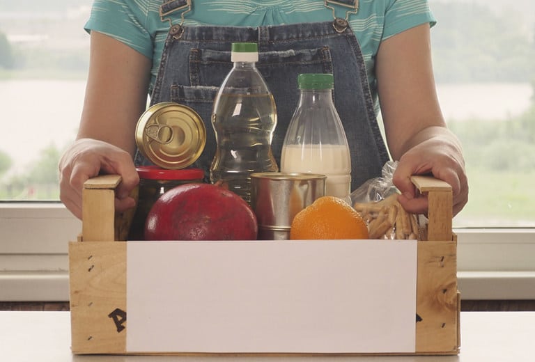 Proyectos Nacionales Persona con camiseta verde y peto vaquero sostiene caja de madera con alimentos básicos: frutas, leche, aceite, conservas y pan. Tonos cálidos y naturales reflejan una acción social solidaria centrada en la entrega de ayuda alimentaria a comunidades vulnerables.