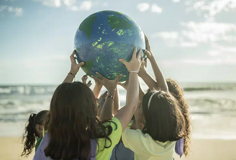 Grupo de niños y niñas en la playa, levantando juntos un globo terráqueo, simbolizando unidad y responsabilidad compartida. El fondo muestra un cielo soleado y el océano, evocando sentimientos de optimismo y compromiso con proyectos que mejoren la calidad de vida de niños y niñas en situación de exclusión social.