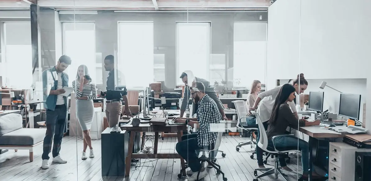 Social Employment Proyect Modern open-plan office with people collaborating at desks and computers, representing acción social in a professional environment. The color palette features neutral whites, grays, browns, and natural light, highlighting teamwork, productivity, and contemporary workspace design.