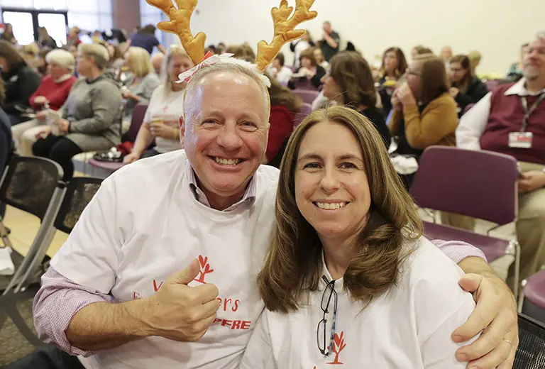 Voluntarios de Fundación MAPFRE sonríen abrazados durante un evento festivo; él lleva una diadema de astas de reno. Ambiente alegre y colores cálidos reflejan una acción social que promueve el espíritu comunitario, la solidaridad y la participación intergeneracional en celebraciones inclusivas.