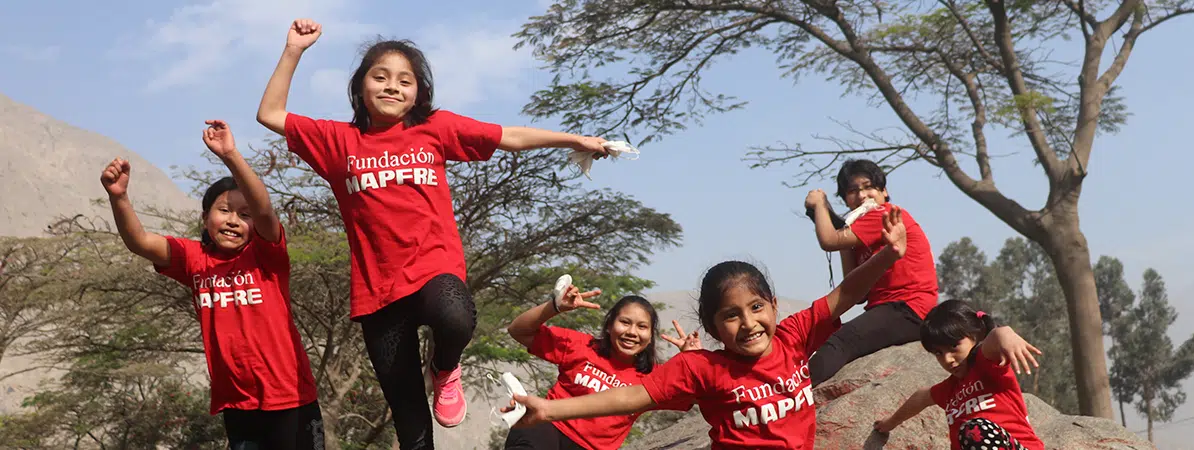 Seis niños y niñas sonriendo y jugando al aire libre, con árboles detrás suyos, todos con camisetas rojas con el logo de "Fundación MAPFRE". Están disfrutando de un momento de alegría en un entorno natural.