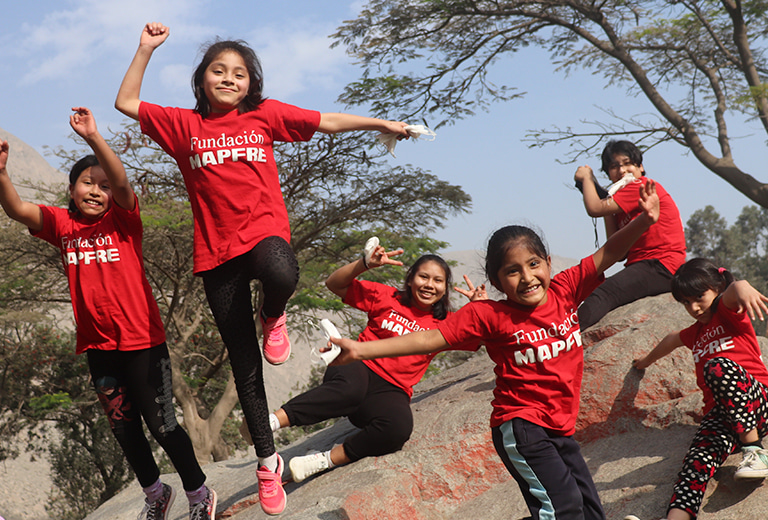 Proyectos Sociales Internacionales Niñas con camisetas rojas de Fundación MAPFRE juegan y saltan sobre rocas al aire libre, en un entorno natural y soleado. La imagen, con tonos vivos y energía positiva, refleja una acción social centrada en la infancia, el ocio saludable y la inclusión.