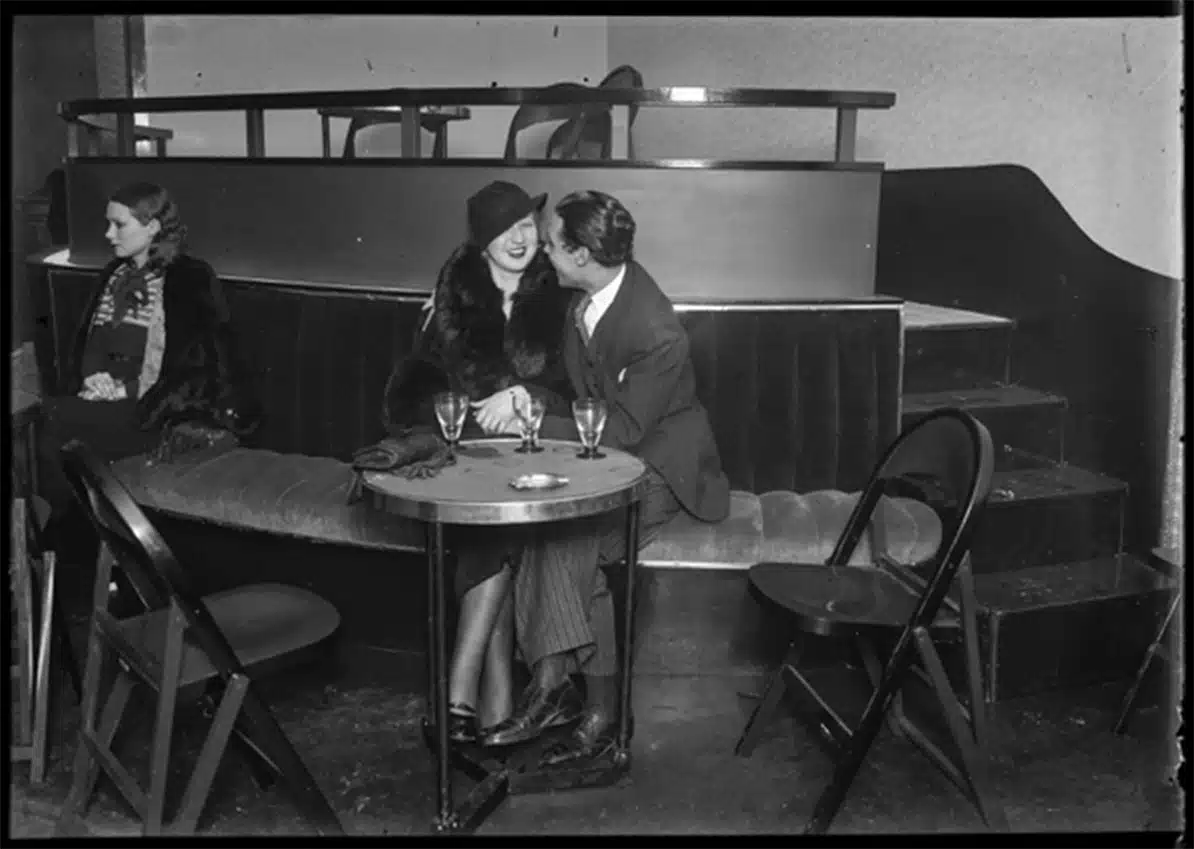 Foto en blanco y negro de una pareja sentada junta en un bar de Barcelona, sonriendo y hablando. Otra mujer está sentada sola cerca. La imagen, titulada «Una pareja en un bar de Barcelona» de Joan Andreu Puig Farran, forma parte de la exposición «Una década de agitación» del KBr.