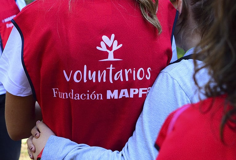 Personas abrazadas por la espalda visten chalecos rojos de voluntarios Fundación MAPFRE, con logotipo blanco sobre fondo vibrante. La imagen transmite unidad y compromiso en una acción social colaborativa que fomenta el voluntariado, la solidaridad y el apoyo comunitario en contextos diversos.