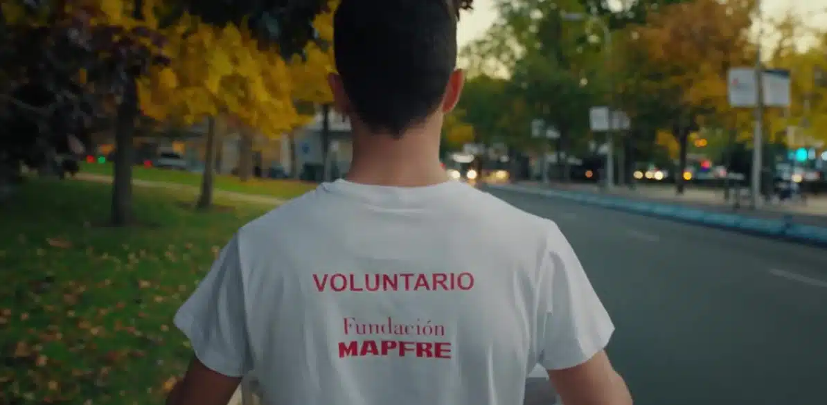 A volunteer wearing a white t-shirt with 'VOLUNTARIO' and Fundación MAPFRE logo walks in an autumn park with yellow and green leaves, promoting acción social and community engagement.