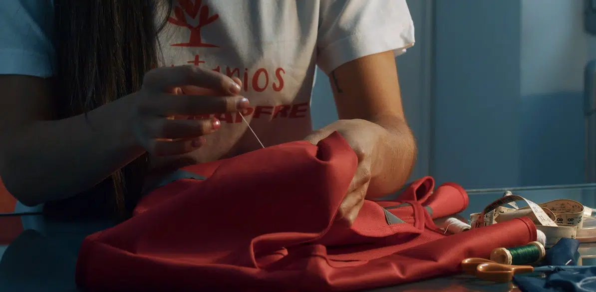 Fundación MAPFRE Persona cosiendo tela roja a mano sobre una mesa con herramientas de costura, como tijeras, hilos y cinta métrica. La escena, en tonos rojos, blancos y azules, refleja un proyecto de acción social enfocado en formación y empoderamiento.